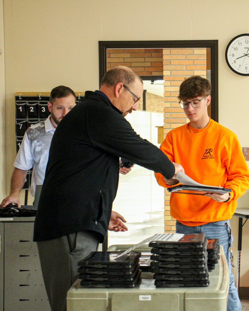 a student in an orange shirt and glasses is handed a Chromebook from a man with glasses and black jacket.