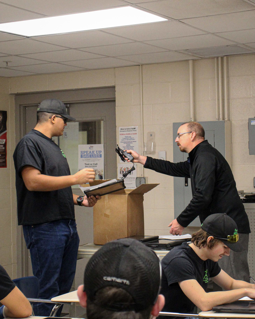 a tall student in an black shirt and hat and glasses is handed a Chromebook from a man with glasses and black jacket.