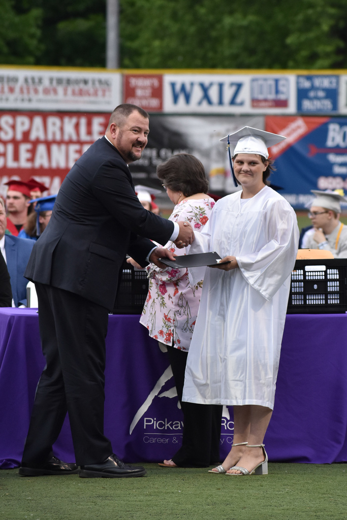 A man in a black suit hands a folder and shakes the hand of a female student in a white graduation robe. 