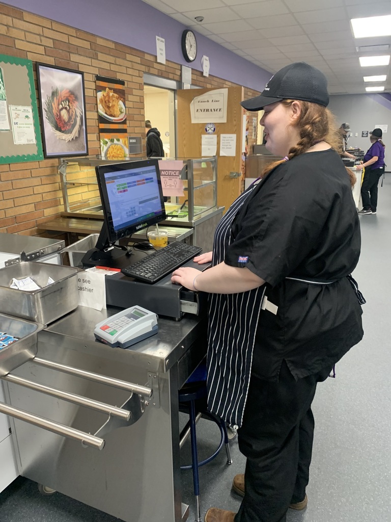 A female student in a black outfit and apron works at a computer.