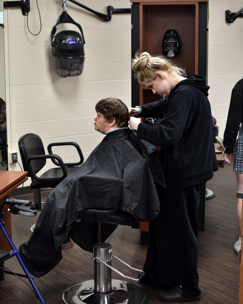 A student in a black sweatshirt and blonde hair cuts the hair of a person in the chair in front of her. 