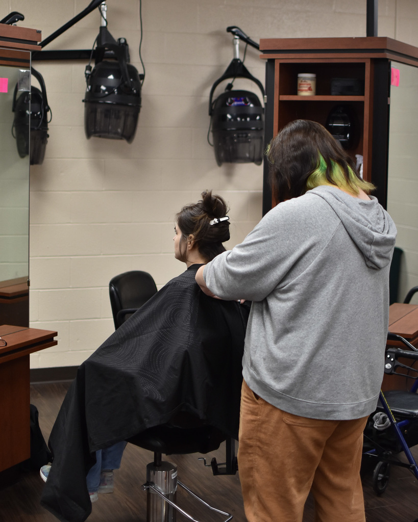 A student in a gray sweatshirt cuts the hair of a person with dark hair.