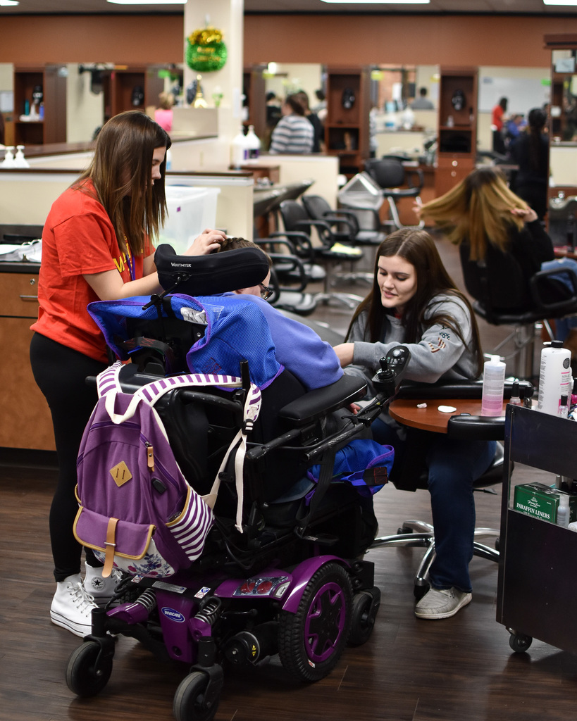 A student in a red shirt braids the hair of a person in a wheelchair while a student in a grey sweatshirt paints their nails.