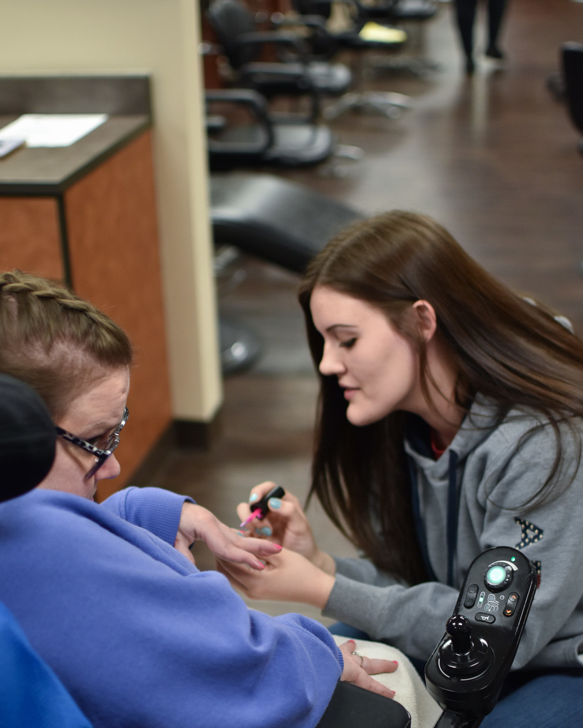 A female student with long brown hair paints the nails of a person in a wheelchair wearing a blue shirt.