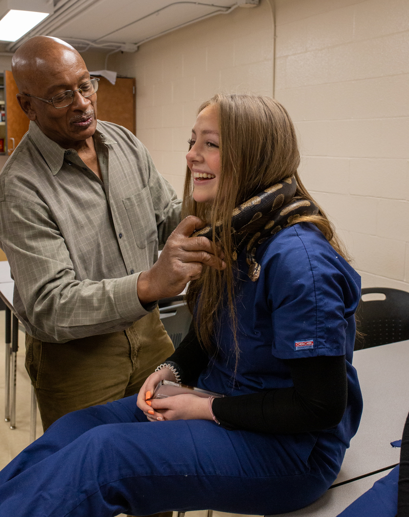 Female student in blue scrubs laughs while a reptile handler assists with a snake around her neck and shoulders. 