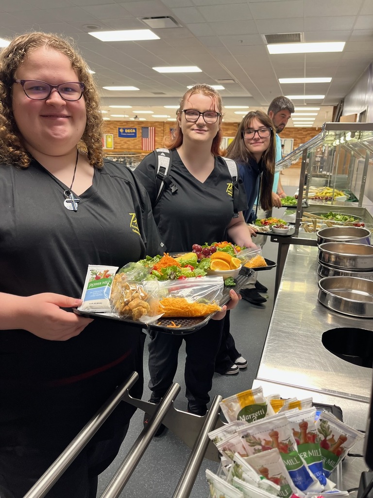 Three female students in gray shorts pose with lunch trays full of salad they created. They stand next to the salad bar in the lunch room. 