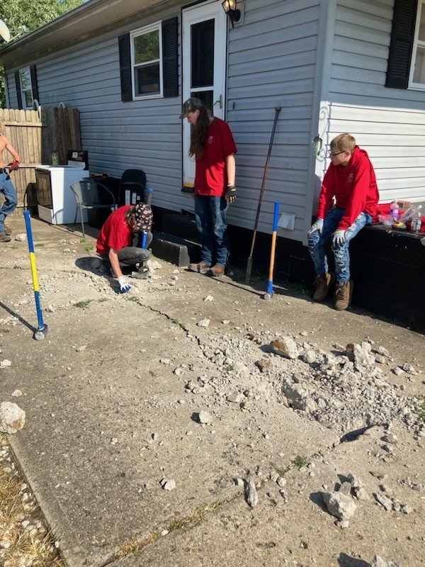 2 boys in red shirts and baseball caps use sledgehammers and other tools to bust concrete from a driveway. Another boy in red sits on a porch nearby.