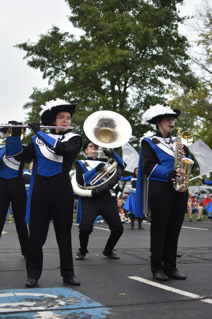 Band members in black, blue and white uniforms with black hates with white feathers preform. One has a flute, one has a tuba and one has a saxophone. 